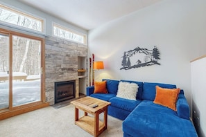Ein Wohnzimmer mit Kamin, einem blauen Sofa, einem Holztisch und Blick auf eine verschneite Landschaft durch große Fenster.