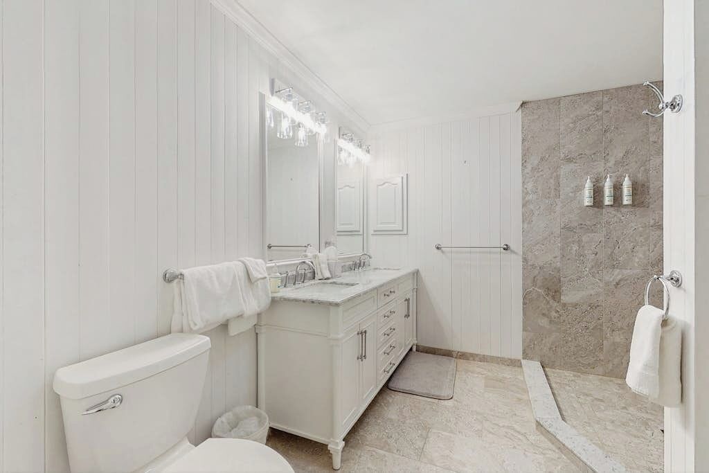A bathroom with a white vanity, a large mirror, a shower area, and marble walls.