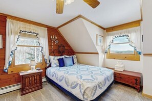 Ein Schlafzimmer mit schrägen Wänden, einem Bett mit gemustertem Überwurf, zwei Fenstern mit Vorhängen, einem Nachttisch und einer Kommode.