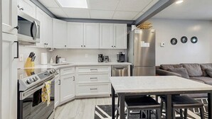 Eine moderne Küche mit weißen Schränken, einer Mikrowelle, einem Herd und einem Kühlschrank. Im Wohnbereich befindet sich ein Esstisch mit Stühlen und ein graues Sofa.