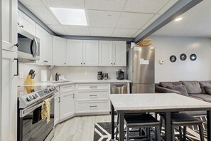 Eine moderne Küche mit weißen Schränken, einer Mikrowelle, einem Herd und einem Kühlschrank. Im Wohnbereich befindet sich ein Esstisch mit Stühlen und ein graues Sofa.