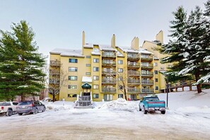 A multi-story yellow building with balconies, a covered entrance, and a parking area with snow. - High-floor resort condo with free WiFi and full kitchen - walk to lifts (Killington)