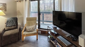 A living room with a sofa, a recliner with a cushion, a flat-screen TV on a wooden stand, and a sliding glass door leading to an outdoor deck. - Resort condo with balcony & shared seasonal amenities - close to skiing (Killington)