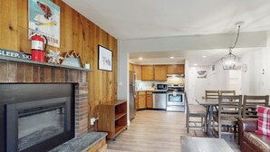A modern living room with a fireplace, wooden walls, a dining area with a table and chairs, and a kitchen in the background. - Resort condo with balcony & shared seasonal amenities - close to skiing (Killington)