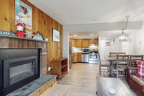 Ein modernes Wohnzimmer mit Kamin, Holzwänden, einem Essbereich mit Tisch und Stühlen sowie einer Küche im Hintergrund.