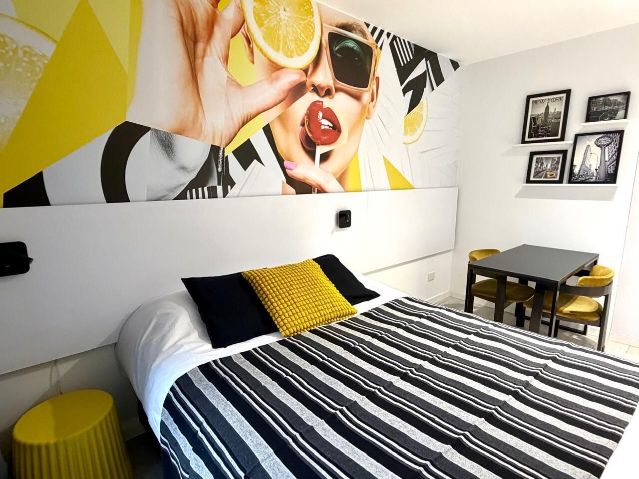 Un dormitorio moderno con una cama a rayas, un almohábil amarillo, un mural blanco y negro en la pared, una mesita con sillas amarillas y cuadros enmarcados en la pared.