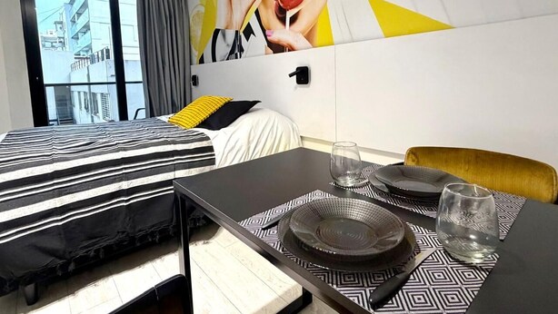 A hotel room with a bed, a dining table set with plates and glasses, a large window, and a wall mural. - CloudBA Fitz Roy Apartments (Buenos Aires)
