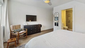 A bedroom with a bed, a desk, a chair, a dresser, a television, and a bathroom visible through an open door. - Comfortable condo w/ mountain & ski views, central a/c, shared patio & BBQ (Waitsfield)