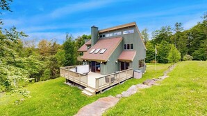 A two-story house with a deck, surrounded by greenery. - 3-story secluded home with private sauna, hot tub, pond on-site (Killington)