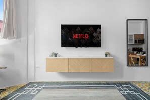 A modern living room with a mounted TV displaying the Netflix logo, a wooden cabinet below, a patterned rug, and a dining area visible in the mirror reflection. - Bellavista Galleria | Old San Juan (San Juan)