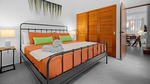 A modern bedroom with a metal bed frame, orange bedding, a wooden wardrobe, and a dining area visible through an open door. - Bellavista Galleria | Old San Juan (San Juan)