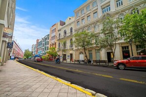 A street lined with multi-story buildings, a sidewalk, and a few parked cars. - Bellavista Galleria | Old San Juan (San Juan)