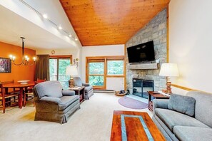 A living room with a stone fireplace, a flat-screen TV mounted above it, a dining area with a wooden table and chairs, and a large window with a view of trees. - Trail Creek 31 | Walk-to-lift condo in Killington with pool, hot tub, gym, sauna (Killington)