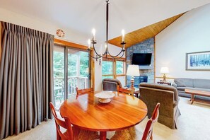 A dining area with a wooden table and chairs, a chandelier, and a television in the background. - Trail Creek 31 | Walk-to-lift condo in Killington with pool, hot tub, gym, sauna (Killington)
