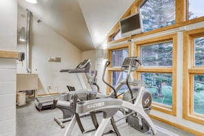 Ein Fitnessraum mit Laufbändern und einem Ergometer, großen Fenstern und einem Fernseher an der Wand.