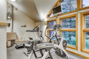 A gym with treadmills and exercise equipment, large windows with a view of trees, and a television mounted on the wall. - Two-story, walk-to-lift condo with indoor pool, hot tub, sauna, gym & game room (Killington)