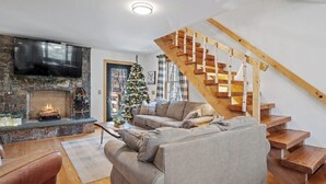 Un soggiorno con camino in pietra, un'ampia televisione a schermo piatto, una scala in legno e un albero di Natale.