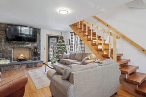 Ein Wohnzimmer mit Kamin aus Stein, einem großen Flachbildfernseher, einer Holztreppe und einem Weihnachtsbaum.