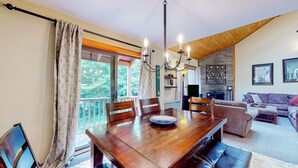 A dining area with a wooden table, chairs, a chandelier, and a view of greenery outside. - Trail Creek 33 | Condo in Killington (Killington)