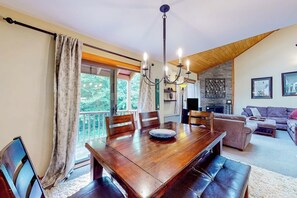 A dining area with a wooden table, chairs, a chandelier, and a view of greenery outside. - Trail Creek 33 | Condo in Killington (Killington)