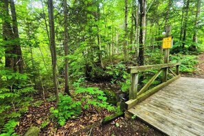 A wooden bridge leading into a forested area with a yellow sign on a post. - Trail Creek 33 | Condo in Killington (Killington)