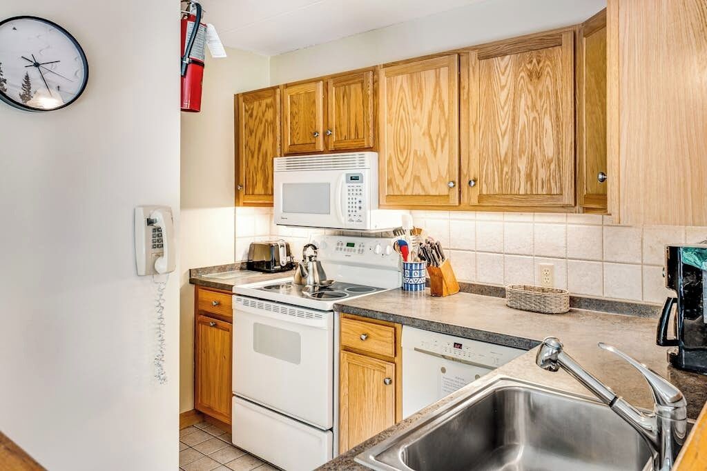 A compact kitchen with wooden cabinets, a microwave, a stove, a sink, and a clock on the wall.