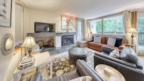 A living room with a fireplace, sofa, ottoman, and a television. - Resort condo with 3 pools, sports courts, & convenient ski access (Warren)