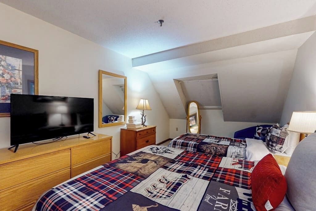 A bedroom with a television, dresser, bed with a plaid bedspread, and a mirror.