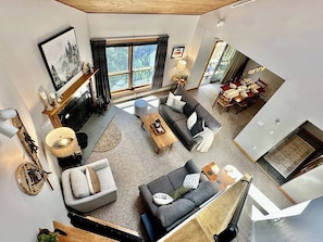 A spacious living area with a sofa, coffee table, and fireplace. There is a dining area with chairs and a table. Large windows provide natural light and a view of the outdoors. - Top-Floor Condo, ski-home trail, w/ fireplace, balcony, w/d, hot tub, pool, wifi (Killington)