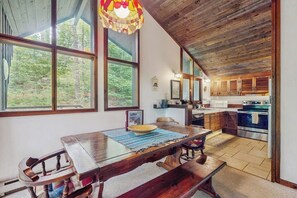 Un interior rústico con una mesa de comedor de madera, ventanas amplias, una cocina con armarios de madera y una lámpara colgante.