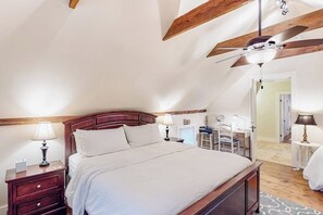 A bedroom with a wooden ceiling, a bed with white bedding, a desk with a chair, and a small table with a lamp.