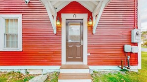 A red wooden house with a brown front door, a small porch, and two wall-mounted lights. - Charming dog-friendly home with private deck, firepit, washer/dryer, & cable (Wallingford)