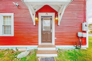 A red wooden house with a brown front door, a small porch, and two wall-mounted lights.