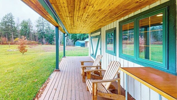 A covered porch with wooden chairs and a table, a grassy yard, and a view of trees and a building in the background. - Cozy house in the woods, riverfront, trail,  fireplace, wifi, w/d, bbq, balcony (Moretown)