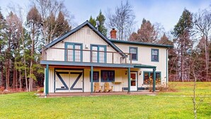 A two-story house with a front porch, a garage, and a small deck. - Cozy house in the woods, riverfront, trail,  fireplace, wifi, w/d, bbq, balcony (Moretown)