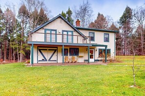 A two-story house with a front porch, a garage, and a small deck. - Cozy house in the woods, riverfront, trail,  fireplace, wifi, w/d, bbq, balcony (Moretown)