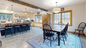A kitchen with a large island, bar stools, a dining table with chairs, pendant lights, and large windows. - Cozy house in the woods, riverfront, trail,  fireplace, wifi, w/d, bbq, balcony (Moretown)