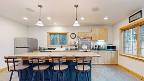 A modern kitchen with a central island, bar stools, wooden cabinets, stainless steel appliances, and a window with a view. - Cozy house in the woods, riverfront, trail,  fireplace, wifi, w/d, bbq, balcony (Moretown)