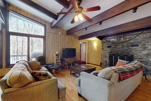 Ein gemütliches Wohnzimmer mit Kamin aus Stein, Holzbalken, einem großen Fenster und einem Fernseher.