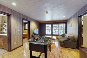 Ein Zimmer mit Holzboden, einem Kicker, einem Sofa, einem Fernseher und einem Fenster mit Blick auf Bäume.