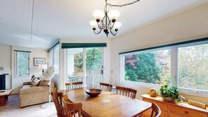 A dining area with a wooden table and chairs, a chandelier, and large windows with a view of trees.