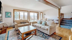 A spacious living room with a sofa, coffee table, and a staircase leading to an upper floor. - Saltbox townhome with views & fireplace - near Quechee Club, golf, & skiing (White River Junction)