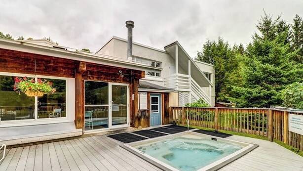 Une terrasse en bois avec un jacuzzi, un patio couvert et un bâtiment doté de grandes fenêtres et d’une cheminée.