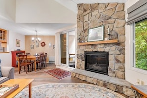 A living room with a stone fireplace, a dining area with chairs and a table, and a view through a glass door. - Convenient Condo with Pool - Mountain Views, Near Town & Slopes (Warren)
