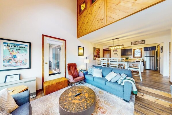 A living room with a blue sofa, a wooden ceiling, a dining area, and a kitchen with a wine cellar sign.