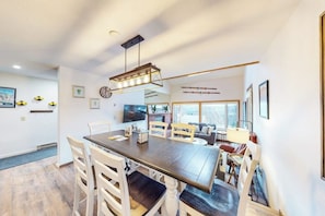 A dining area with a long table, chairs, a hanging light fixture, and a television on the wall.