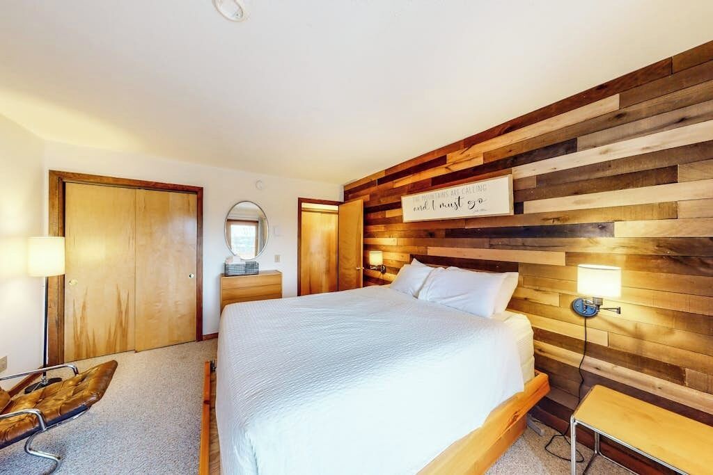 A bedroom with a wooden headboard, a bed with white bedding, a bedside table, a chair, and a mirror.