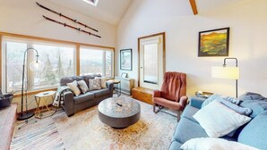A spacious living room with large windows, a sofa, a chair, a coffee table, and skis on the wall.