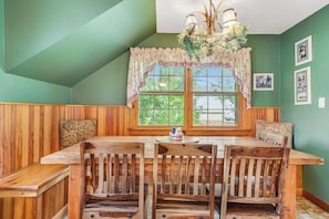 Un coin repas avec une table et des chaises en bois, une fenêtre ornée d’un rideau à motifs floraux et un lustre.