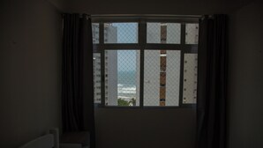 A room with a bed, a window with curtains, and a view of buildings and a body of water. - Florida in Guarujá. Cozy apartment with ocean view. Pets allowed! (Guarujá)